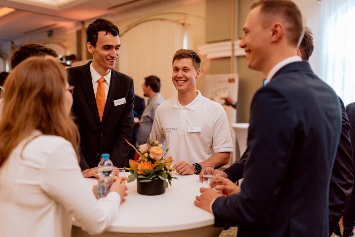 A group of six formally dressed people gather around a high table at an indoor event, captured in this stunning event photography. They are engaged in conversation and smiling, there is a floral centerpiece on the table, and several people are holding bottles or glasses of water. 