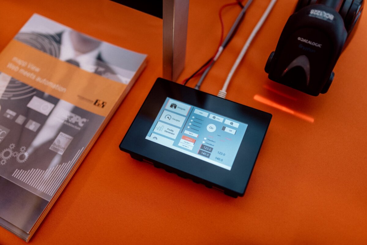 A touch control panel displaying various settings and options is placed on an orange surface. On the left is a brochure with a partial image of a person in a suit, and on the right is a barcode scanner illuminated by a red light. This setup captures the essence of event photography.  