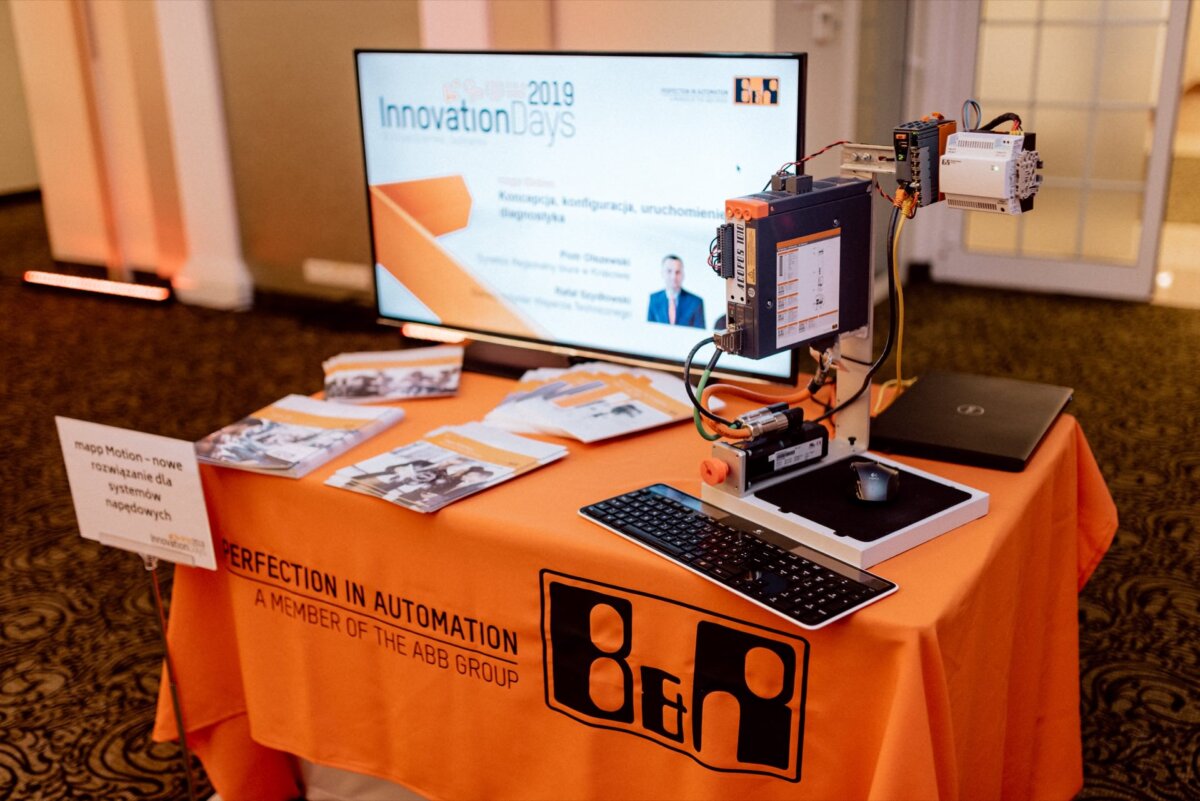 A display table set up at the event shows a computer monitor and keyboard, flyers and brochures, and a robot arm setup. The table is covered with an orange tablecloth with the words "PERFECTION IN AUTOMATION, MEMBER OF THE ABB GROUP." In the background you can see a door and a standing column - perfect for event photography.  