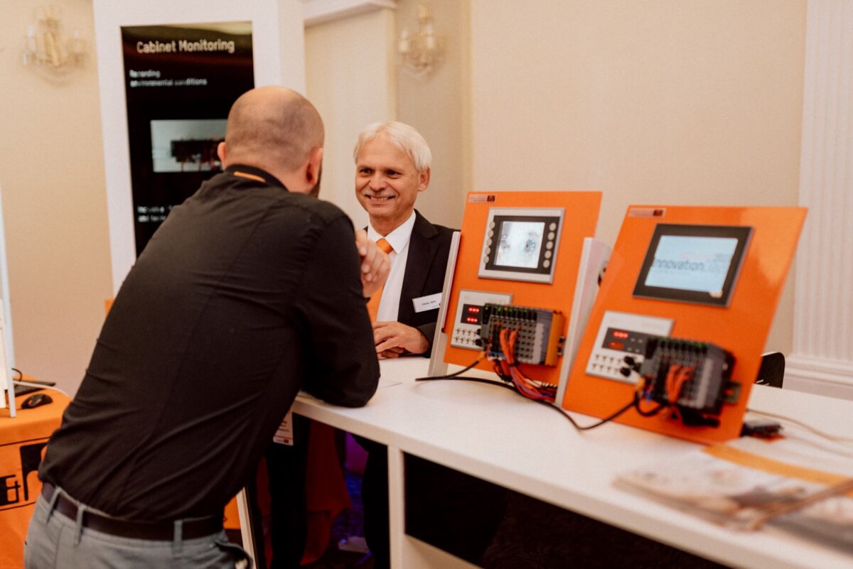 Two men are having a conversation at an exhibition stand. A man with gray hair stands behind the desk, facing the other man, who is bald and standing with his back to the camera. The booth has orange electronic equipment and a screen that captures the perfect moment for event photography.  