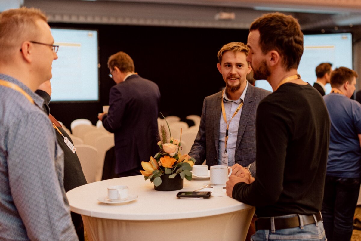 A group of people, mostly men, are standing around a high table talking in conference. Some are holding cups of coffee, and one of the men is gesturing as he speaks. In the background you can see screens and other participants. A flower arrangement adorns the table, perfectly captured by event photographer Warsaw.   