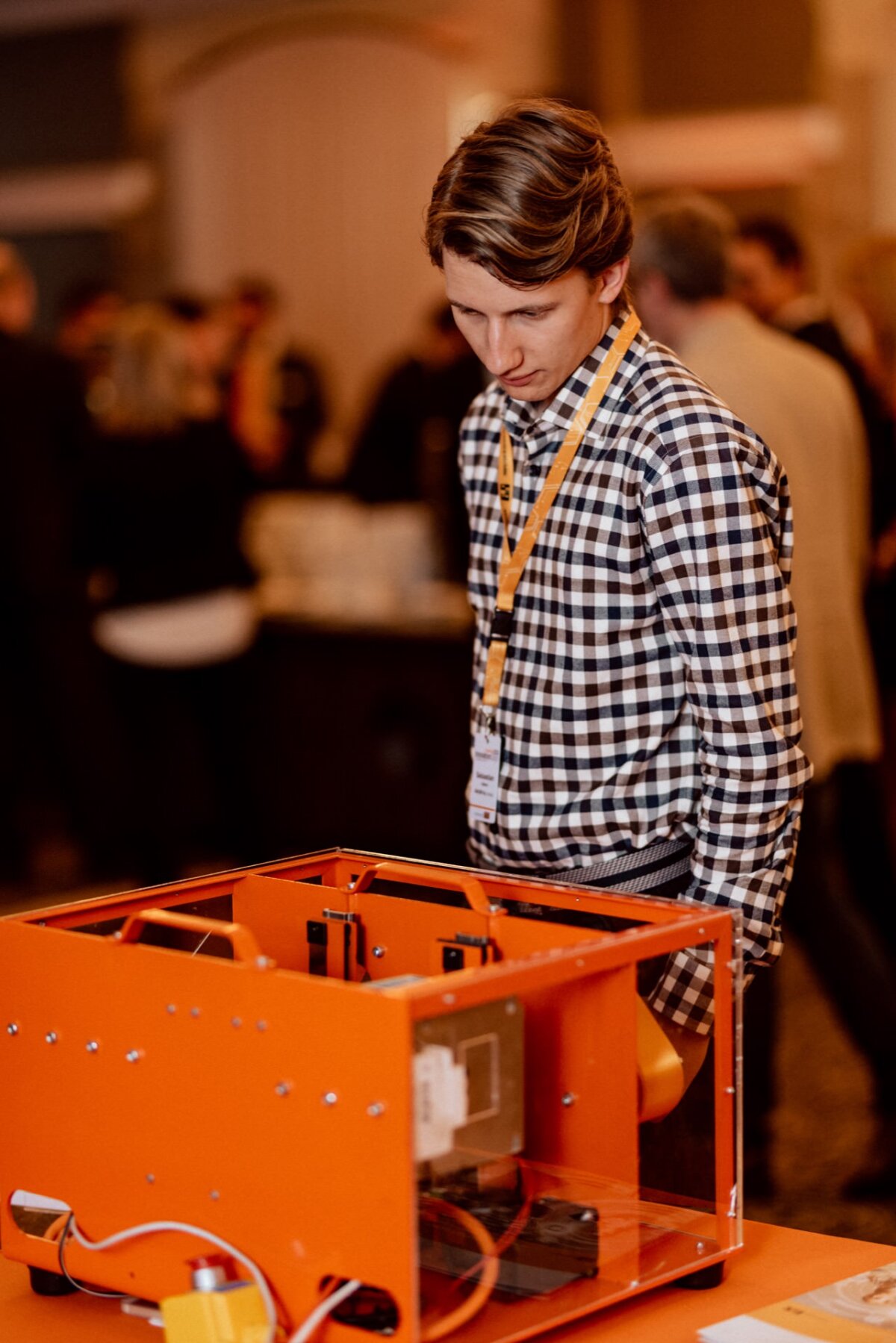 A person wearing a plaid shirt and yellow lanyard looks at an orange electronic device lying on a table. The background is a blur with several other people, reflecting the hustle and bustle often seen in event photo coverage, suggesting a busy setting for an event or conference. 