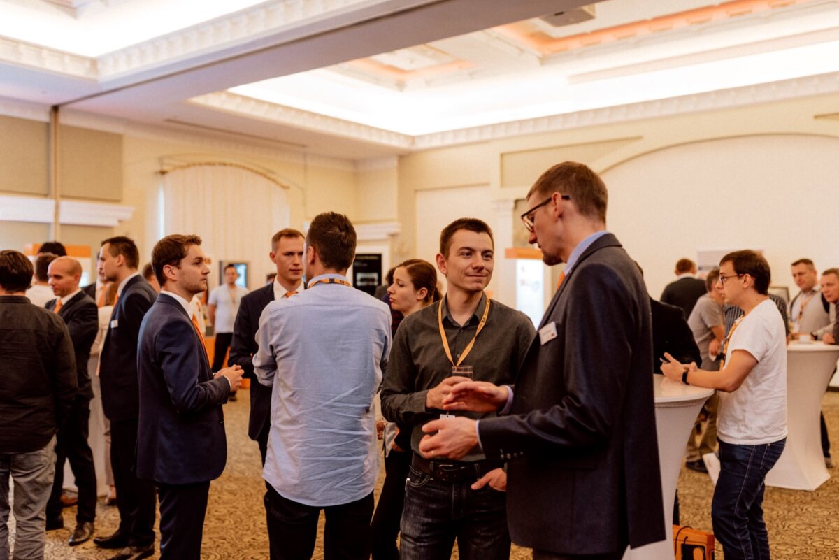 A group of people, mostly men, gather in a well-lit room to attend a professional networking event. Some are busy talking, while others stand in the background. The room has a formal atmosphere with high ceilings and round tables - perfect for capturing photo-reports of events.  