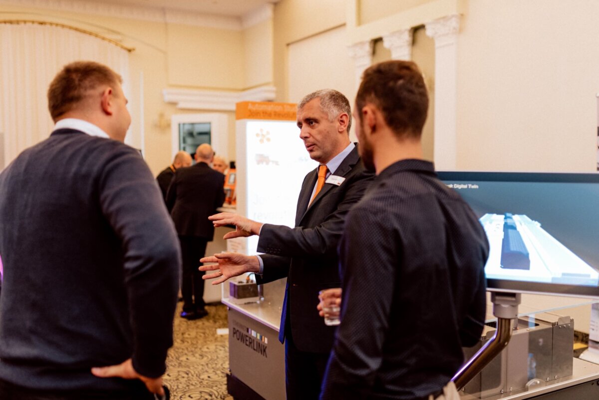 Three men in business attire are talking at a technology conference. One of the men, gesturing with his hands, seems to be explaining something. In the background, a presentation screen and a rack of technology equipment point to a well-documented event photo by an event photographer warsaw.  