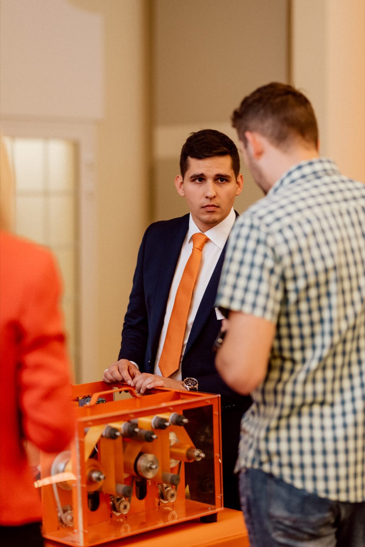 A man dressed in a suit and orange tie is standing behind an orange mechanical parts display table and having a conversation with a casually dressed man. On the left side of the photo, another person with blond hair and an orange jacket is partially visible, beautifully captured in this event photo. 