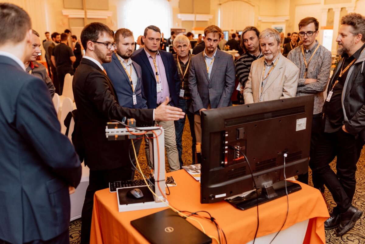 A group of men stand around an orange-covered table and listen to a person in a black suit explain something on a monitor. Behind them are white chairs and other people, which captures the essence of the conference setting through event photography in this detailed photo essay. 