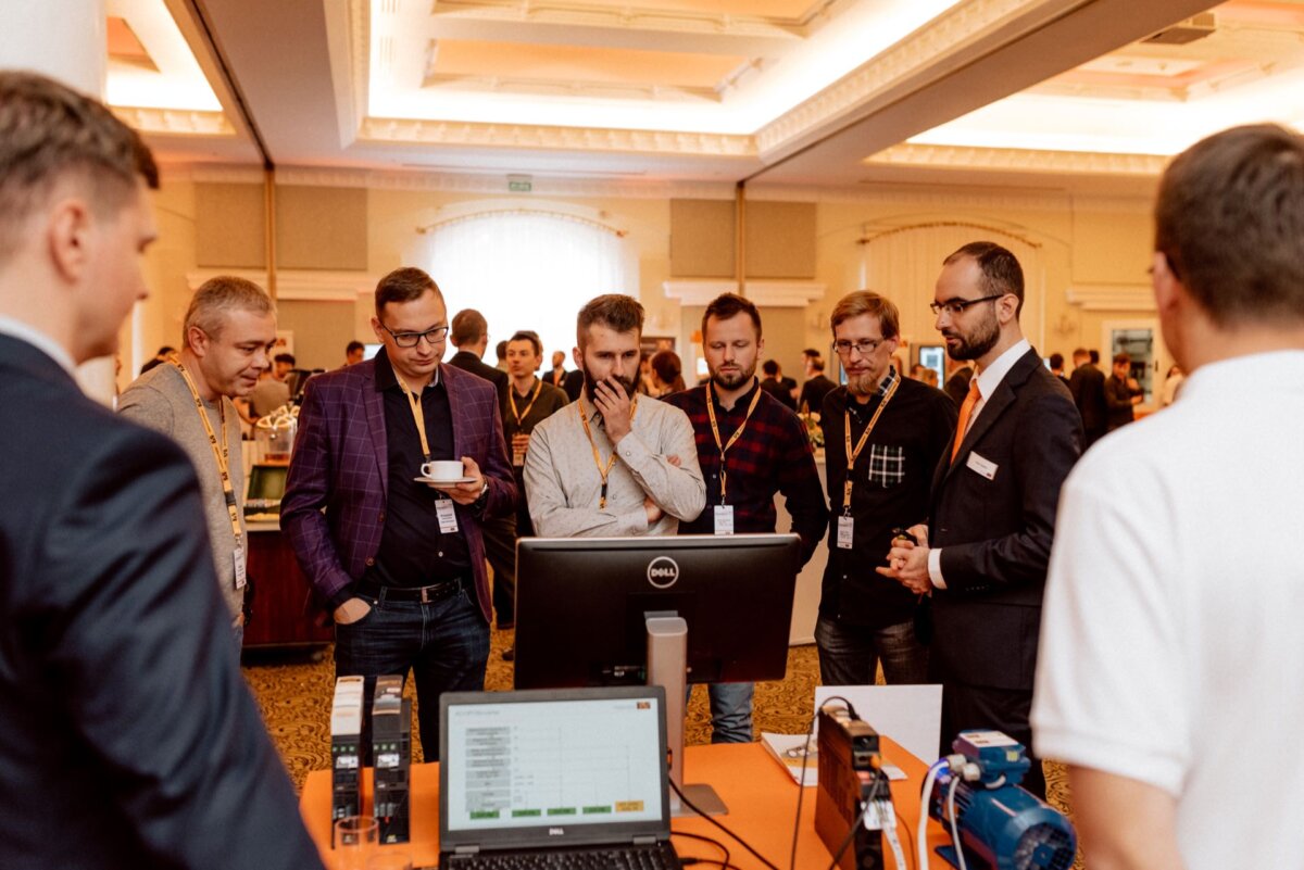 A group of professionally dressed men stand around an exhibit table, carrying on a conversation and looking at equipment at a conference or trade show. One man holds a cup of coffee. A computer monitor and several pieces of equipment lie on the table, captured by event photography so that every moment is professionally documented.  