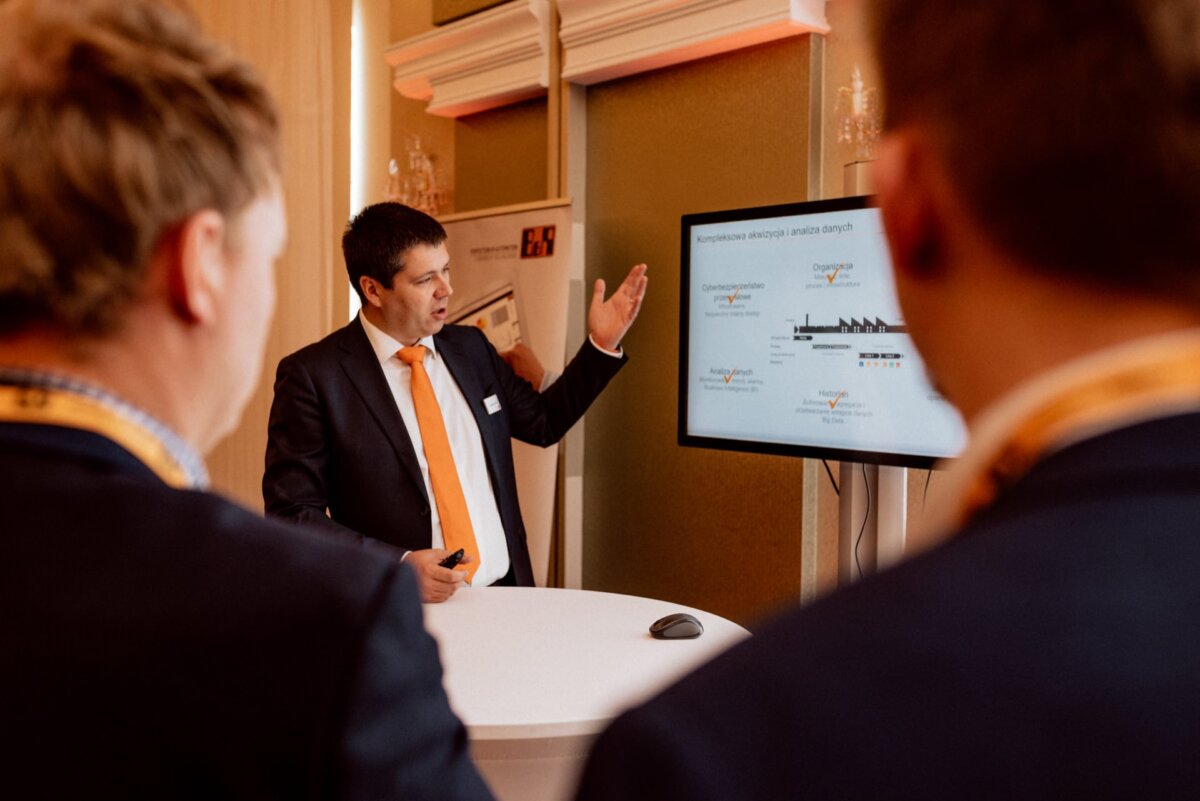 A man in a suit and orange tie gestures during an office presentation. He stands in front of a large screen displaying complex data visualizations, while two other people in suits watch him. A lanyard hangs around the speaker's neck; it's a fascinating snapshot of events.  