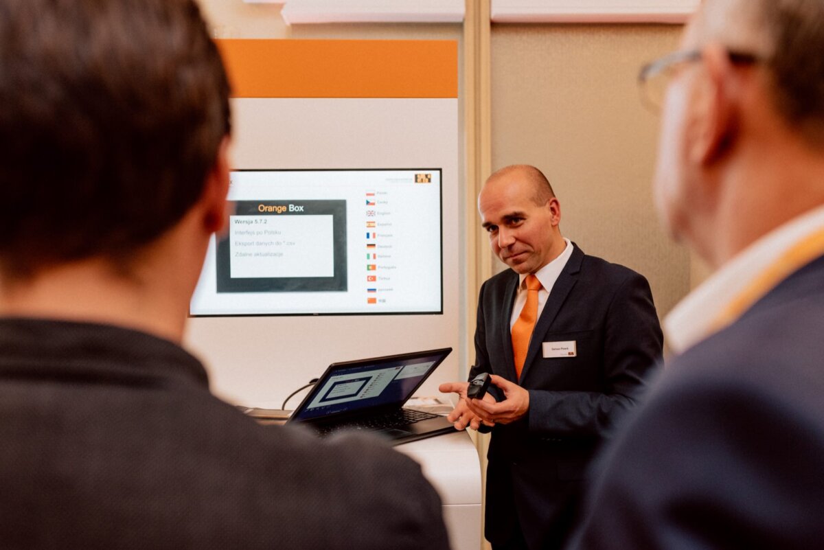 A man in a suit and tie is giving a presentation to two people. He is standing next to a laptop and screen displaying a presentation entitled "Orange Box." The image, ideal for event photography, shows both the focused expression of the presenter and the attentive faces of the audience.  