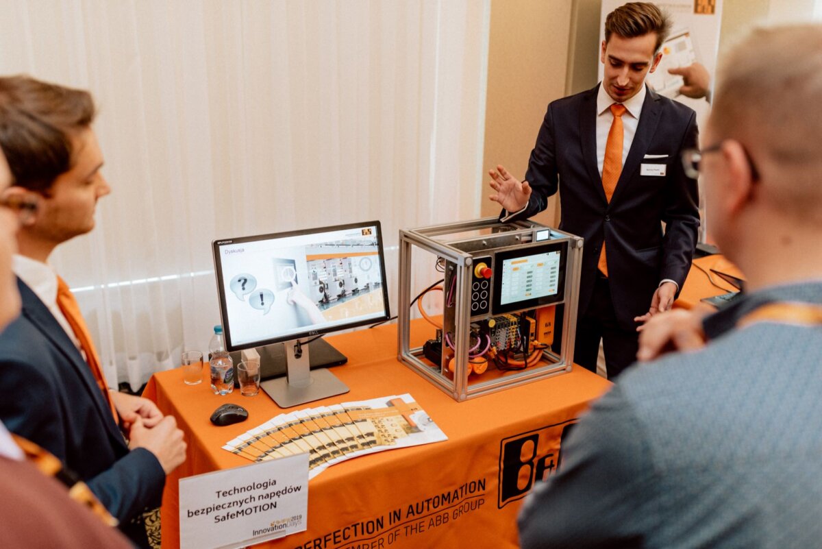 A man in a suit and orange tie demonstrates a piece of automation technology to a small group. The set includes a monitor displaying diagrams and a device standing next to it on a table. A sign on the table read: "SeMOTION transportation technology." Event photography captures every moment.   