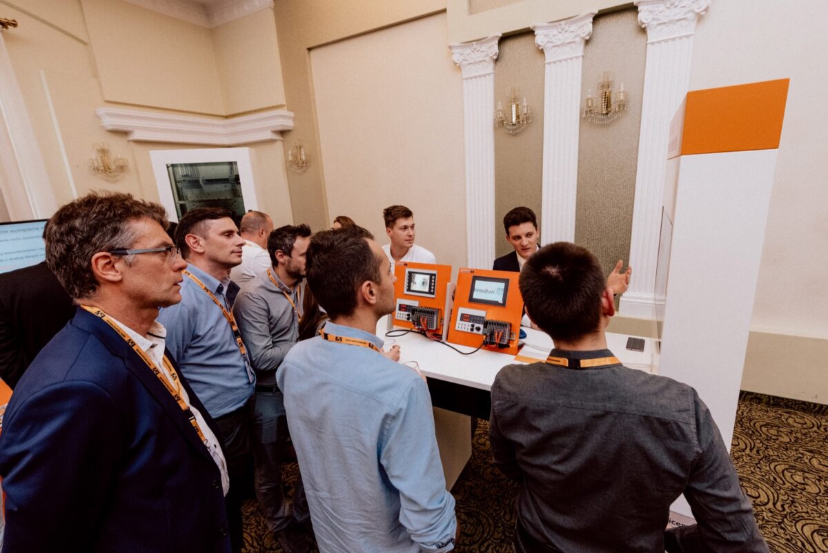 A group of people, mostly men, gather around a display with electronic equipment in a formal setting, such as a conference or exhibition. The equipment has screens and buttons, and one man explains its functions to the others. This moment makes for an ideal photo-reportage of events.  