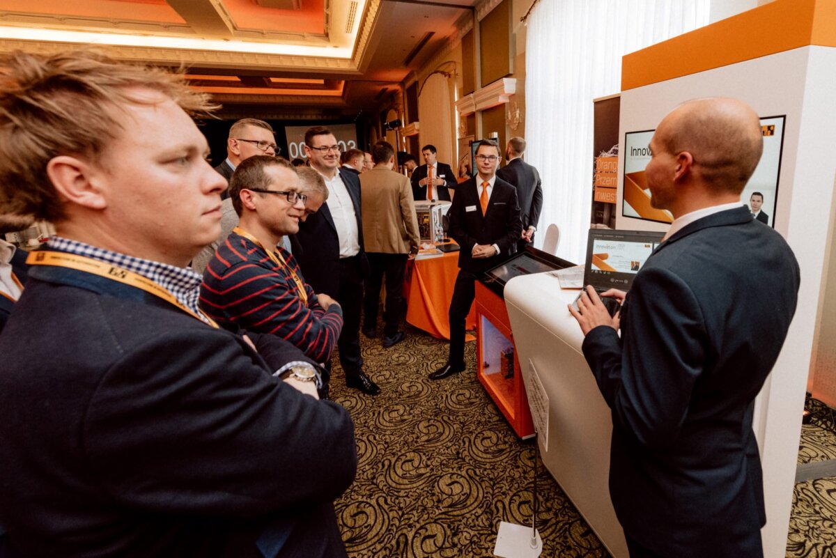 A group of people gathers at a trade show or conference booth. One man, with his back turned to the camera, is giving a presentation or demonstration. Others, dressed in business attire, listen attentively. Booth displays and promotional materials can be seen in this event photograph.   