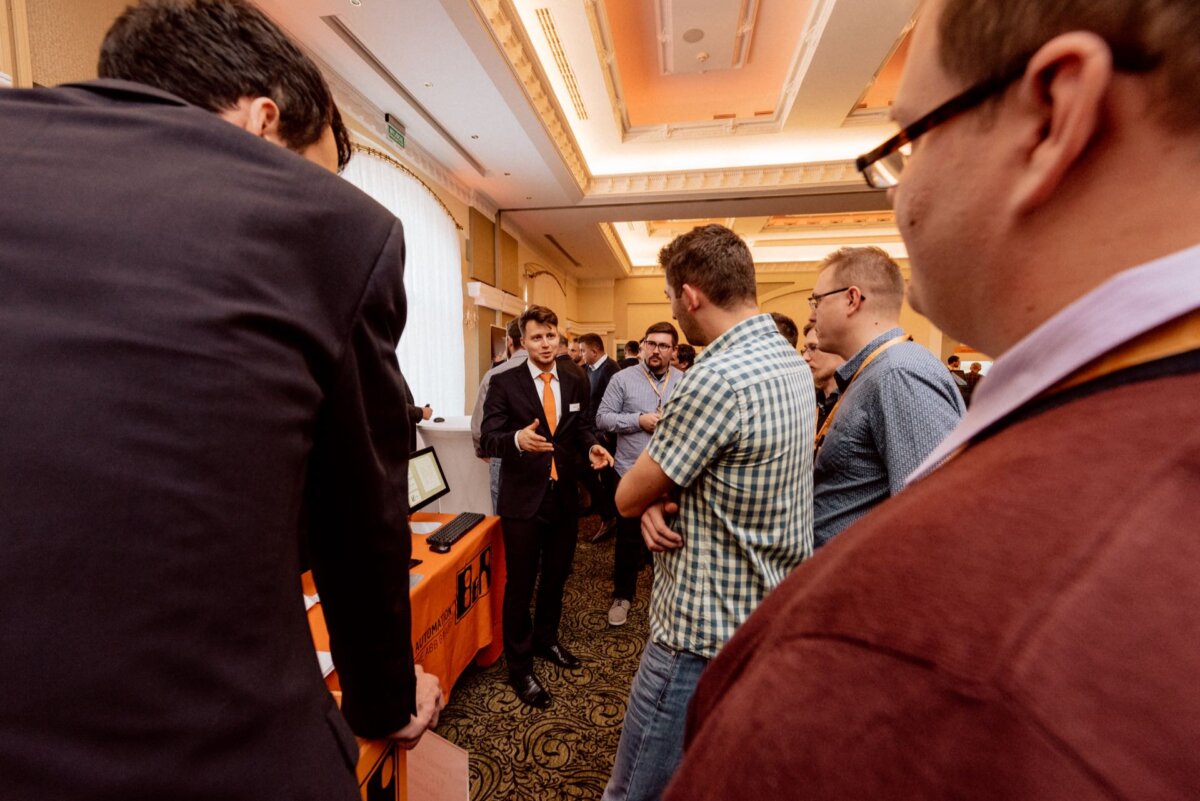 A group of people are gathered in the room, immersed in conversation. One of the men, standing at a table with electronic devices, seems to be explaining something. The others listen attentively. The scenery is reminiscent of an event taking place in a room with warm lighting and ornate ceiling details - perfect for photo coverage of events.   