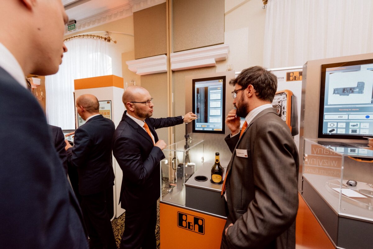 A group of people in formal attire gather around a digital exhibit at a trade show or technology conference. One man points to a touchscreen with a gesture while the others watch and carry on a conversation. The room, ideal for event photography, is well lit, with bright walls and a carpeted floor.  