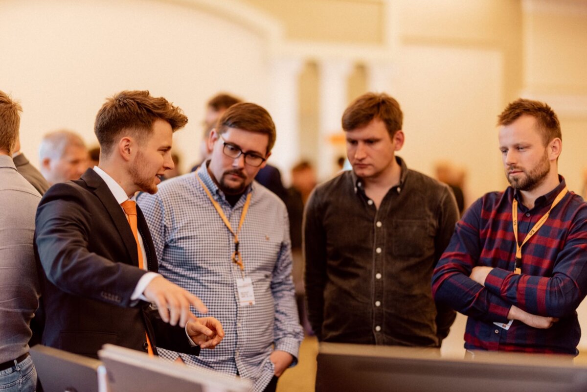 A group of four men have gathered in a room immersed in serious discussion. One man in a suit with an orange tie gestures as he talks to the others, dressed casually. They seem focused on the subject, perhaps at a conference or meeting, captured as part of an event photo shoot (event photography).  