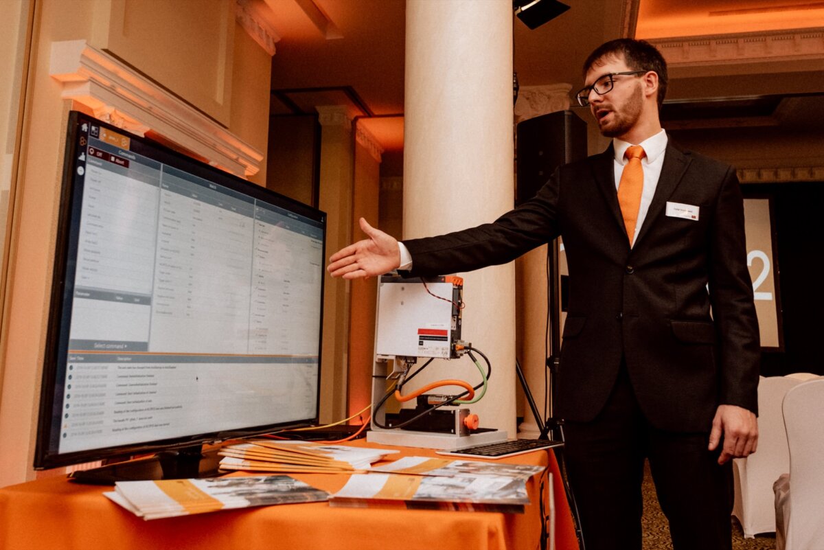 A man in a suit and orange tie stands next to a computer monitor displaying data, extends his hand toward the screen and explains something. The set includes several cables and brochures on an orange-decorated table, perfect for an event photo essay prepared by any event photographer Warsaw. 