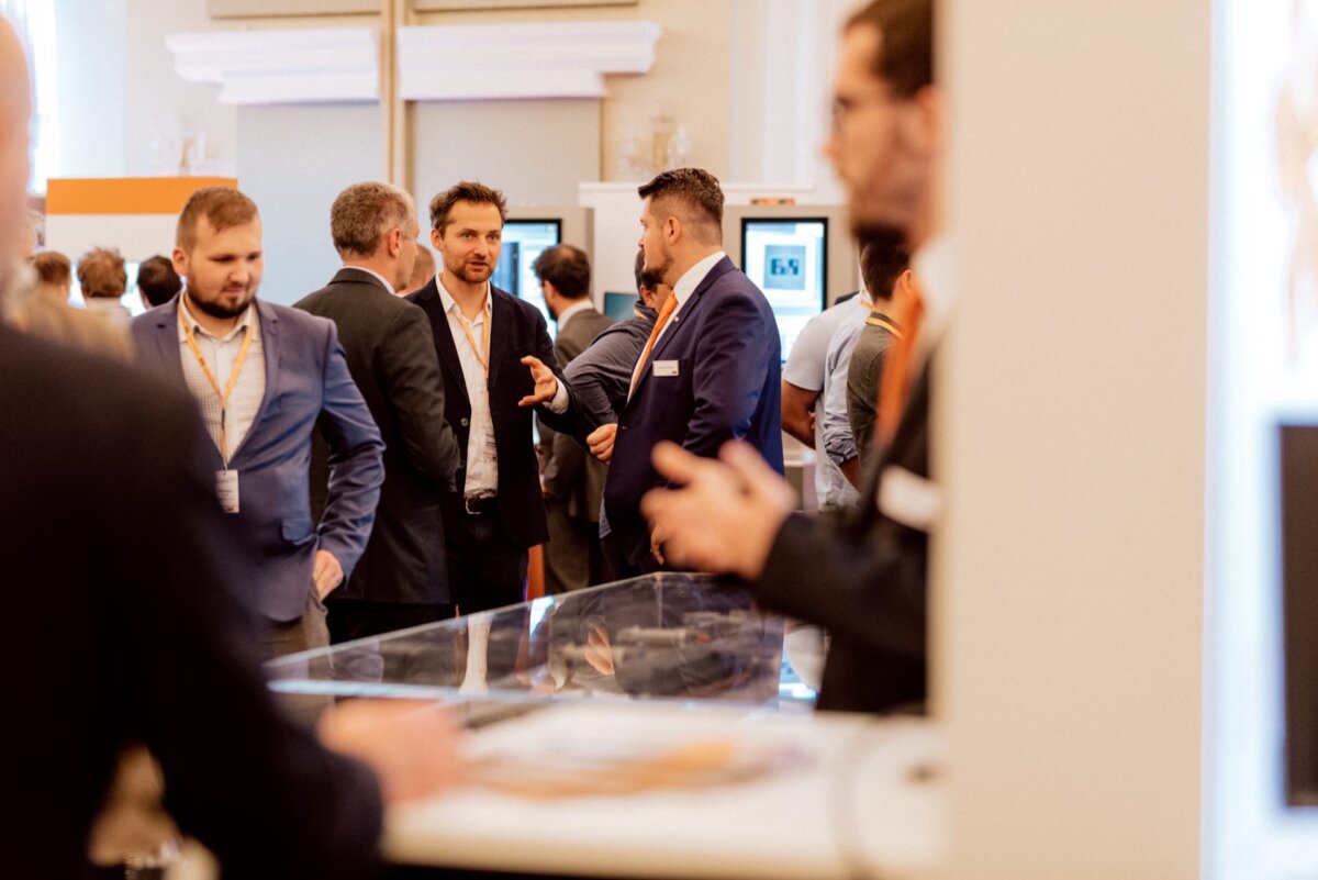 A group of people dressed in business attire are chatting at a networking event or conference. Some stand in small clusters, others walk by. The background depicts booths and displays, suggesting a trade show or professional meeting - perfect for event photography or photo coverage.  