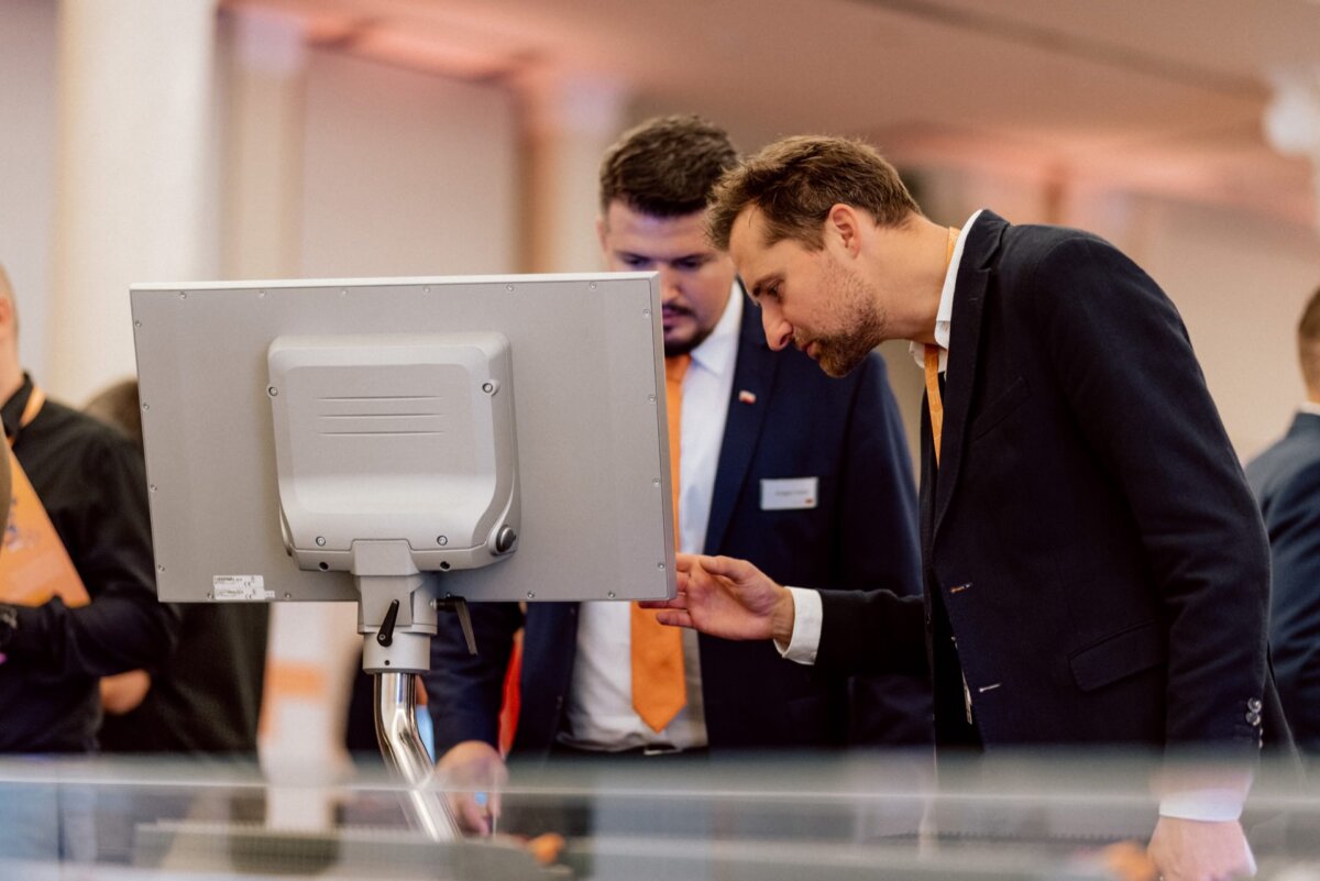 Two men in suits and orange ties stand in front of a large monitor. One man leans over, pointing at the screen, while the other looks on, slightly behind him. The scene captures a moment typical of photo coverage of events in a professional or corporate environment.  