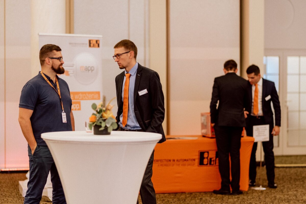 A man in a blue polo shirt and a man in a suit with an orange tie are talking at a high table. In the background, two men in suits with orange ties stand at a booth decorated with orange tablecloths and display materials. A banner with the words "B&amp;R" and "mapp." captures the essence of event photography.