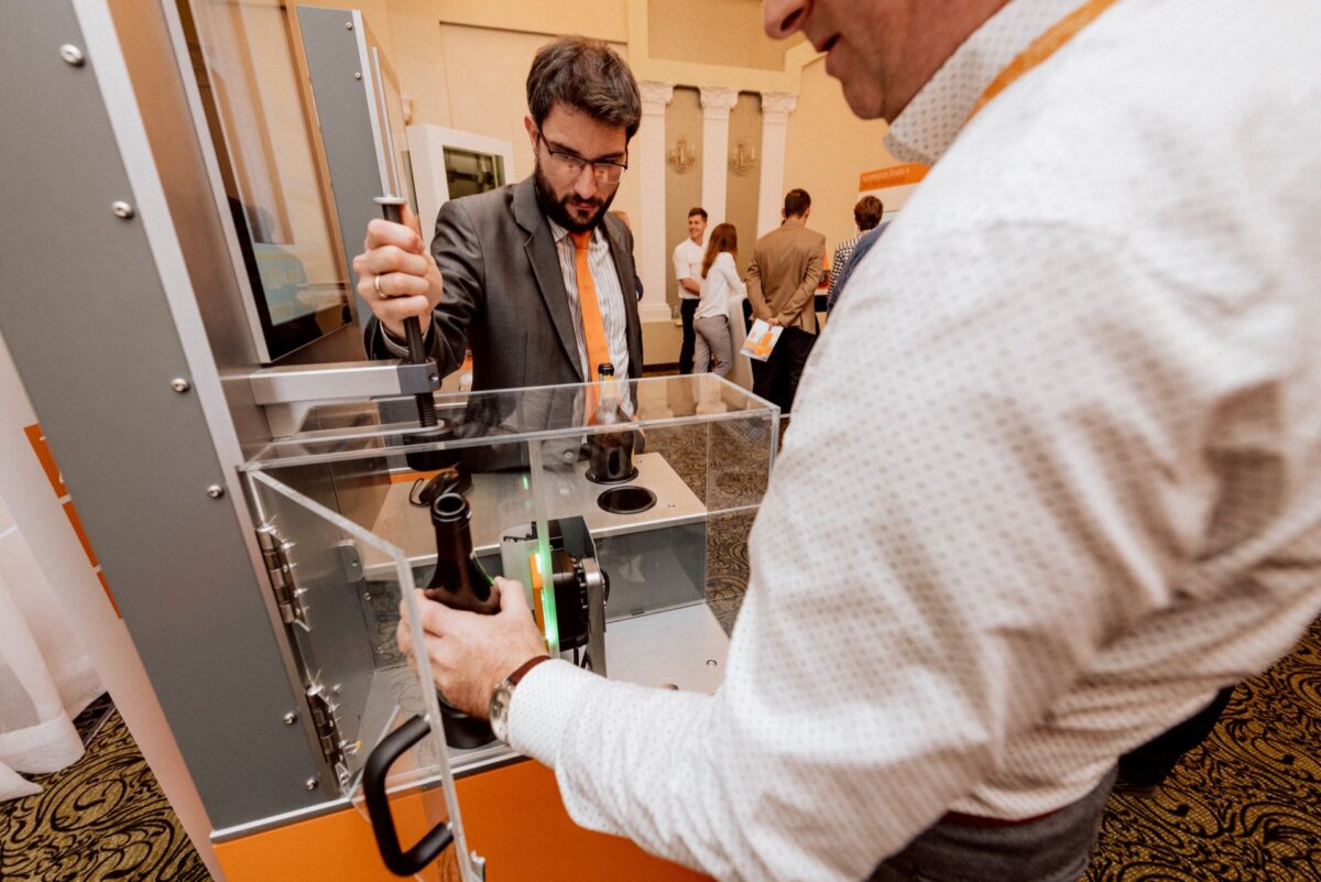 A man in a white shirt operates a machine in a clear glass case, while another man in a suit and tie watches. They are in a room, probably at a trade show or technical demonstration with other people and booths in the background, captured as part of a photo essay of the event. 