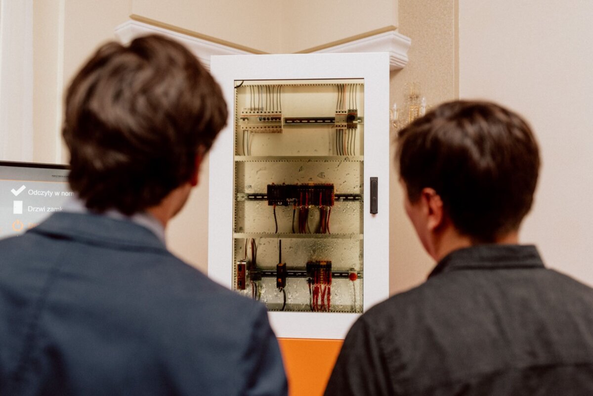 Two dark-haired people, one in a suit and the other in a dark jacket, examine an open electrical panel filled with various wires and components. This photo essay of the event captures them in a detailed interior scene, perfectly captured by the event photographer warsaw. 