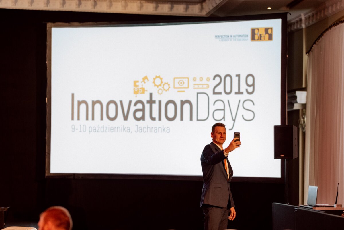 A person in a suit stands on the stage and takes a selfie with his phone. Behind them is a large screen displaying "Innovation Days 2019" and event details in Polish, including dates and location. The venue resembles a conference room, ideal for capturing photo coverage of events.  