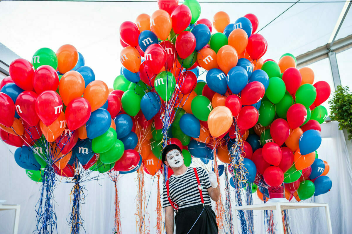 Mime is standing in front of a large collection of colorful balloons, mostly red, blue, green and orange, decorated with the letter "m." The mime is dressed in a black and white outfit, wearing a striped shirt and suspenders, which gives him a playful expression. The scene was captured by Marcin Krokowski, an acclaimed event photographer Warsaw.  