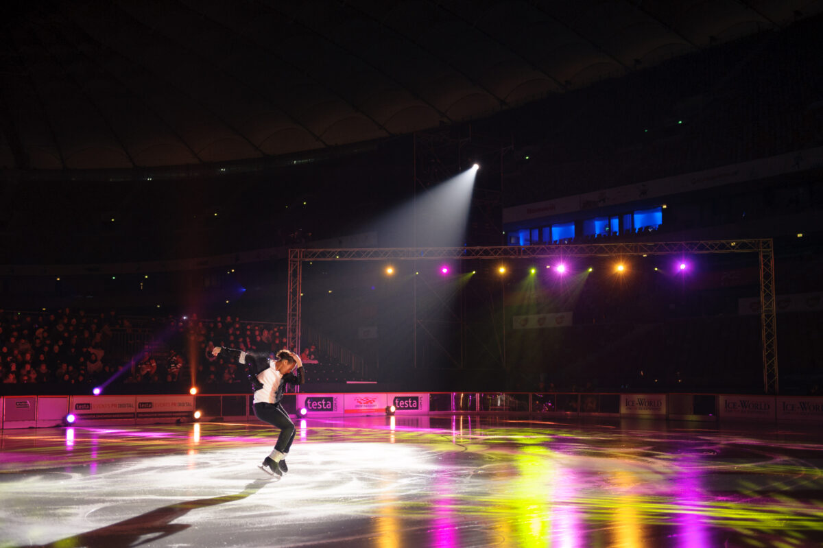 A figure skater in a white shirt and black pants performs on an illuminated ice rink under the spotlight. The rink, beautifully decorated with colorful lights and banners, adds to the enchanting atmosphere. Marcin Krokowski captured the magic of the event, showcasing his talent as an event photographer warszawa.  