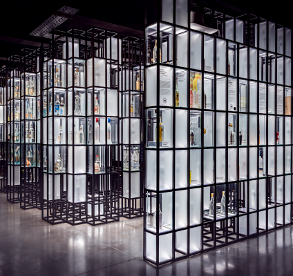 The modern display consists of a number of tall, illuminated glass cases arranged in rows, displaying a variety of bottles and objects. Structural metal frames add a sleek, contemporary touch to the display. An event photographer captures the illuminated display cases reflected on the glossy surrounding floor.  