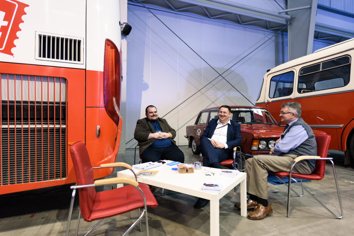 Three men sit around a small white table in a garage-like setting, immersed in conversation and surrounded by vintage buses. On the table are various objects, including papers and cups. The table is surrounded by red chairs, and a section of a red-and-white bus can be seen on the left - a captivating photo-reportage of the events.  