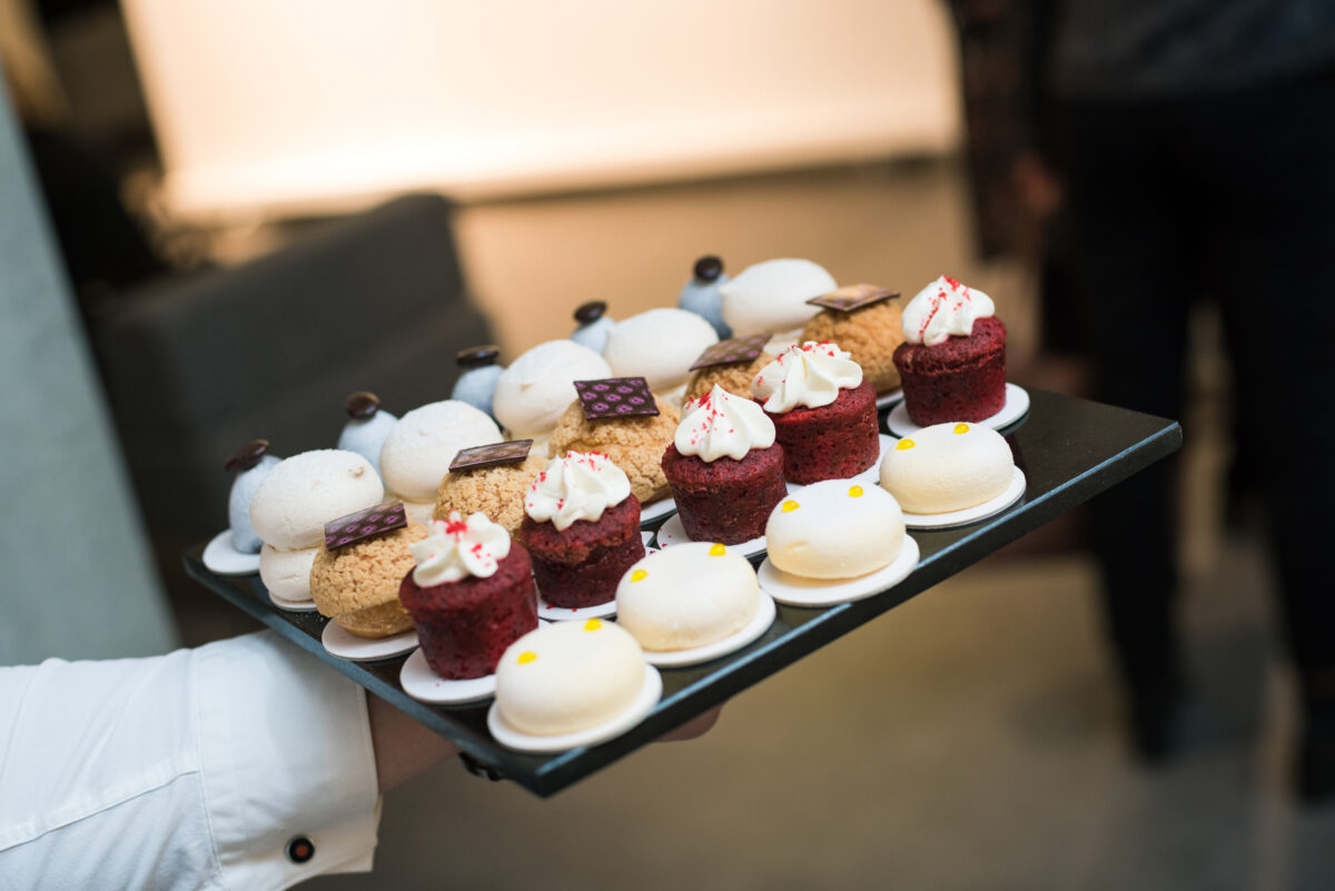 A tray with a variety of desserts, including white macaroons, small red velvet cupcakes covered in white icing, round cakes filled with cream and purple treats. The tray is held by someone wearing a white shirt. This photo shows a delightful moment from a recent event photographer in Warsaw. The background is slightly blurred.   