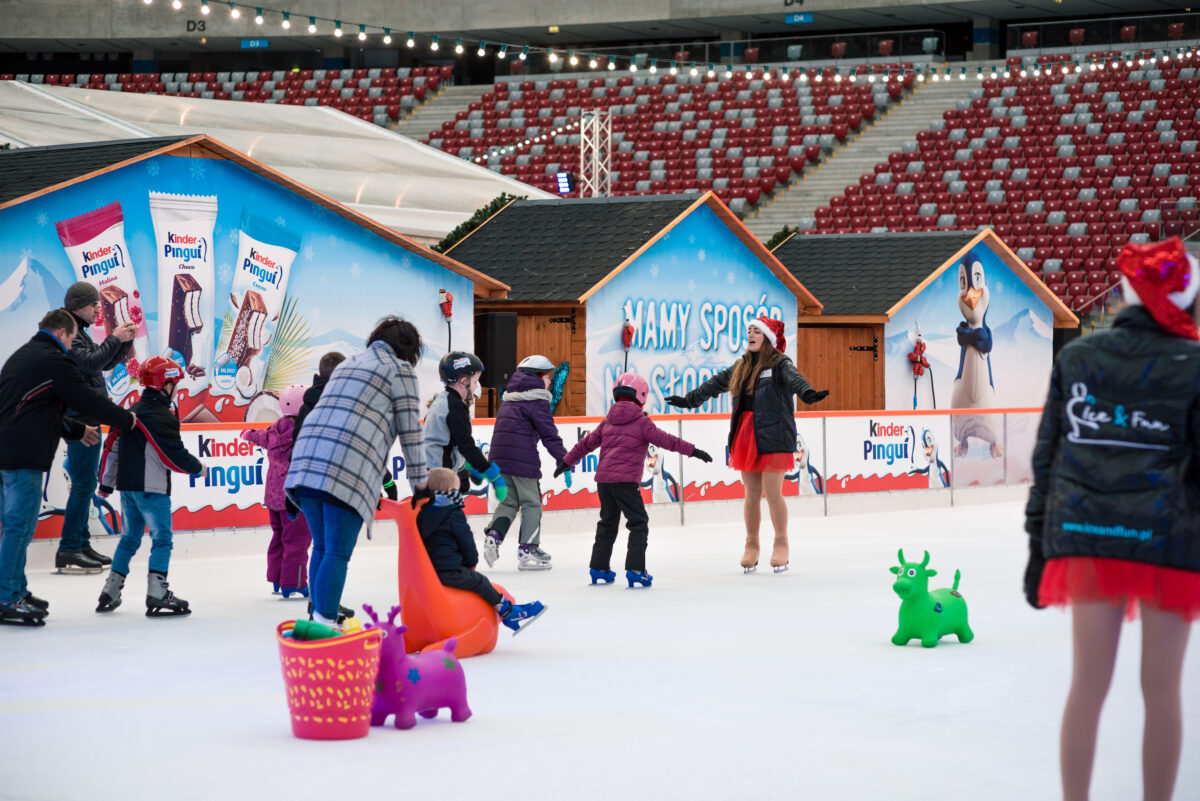 A group of people skate on an outdoor ice rink. Some skaters hold up colorful plastic skating aids in the shape of penguins and other animals. In the background are holiday stalls and advertising banners. The atmosphere is lively and joyful, perfect for an event photographer capturing the moment.   