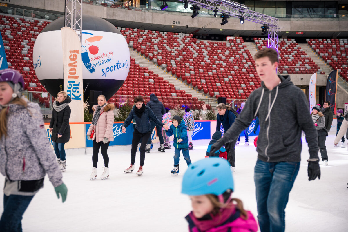 A group of people, including children and adults, skate on an indoor ice rink with red stadium stands in the background. Some are wearing helmets and colorful winter clothes. Above their heads, a large balloon with the logo and an arch with Christmas lights are visible, capturing the lively atmosphere of event photographer Marcin Krokowski.  