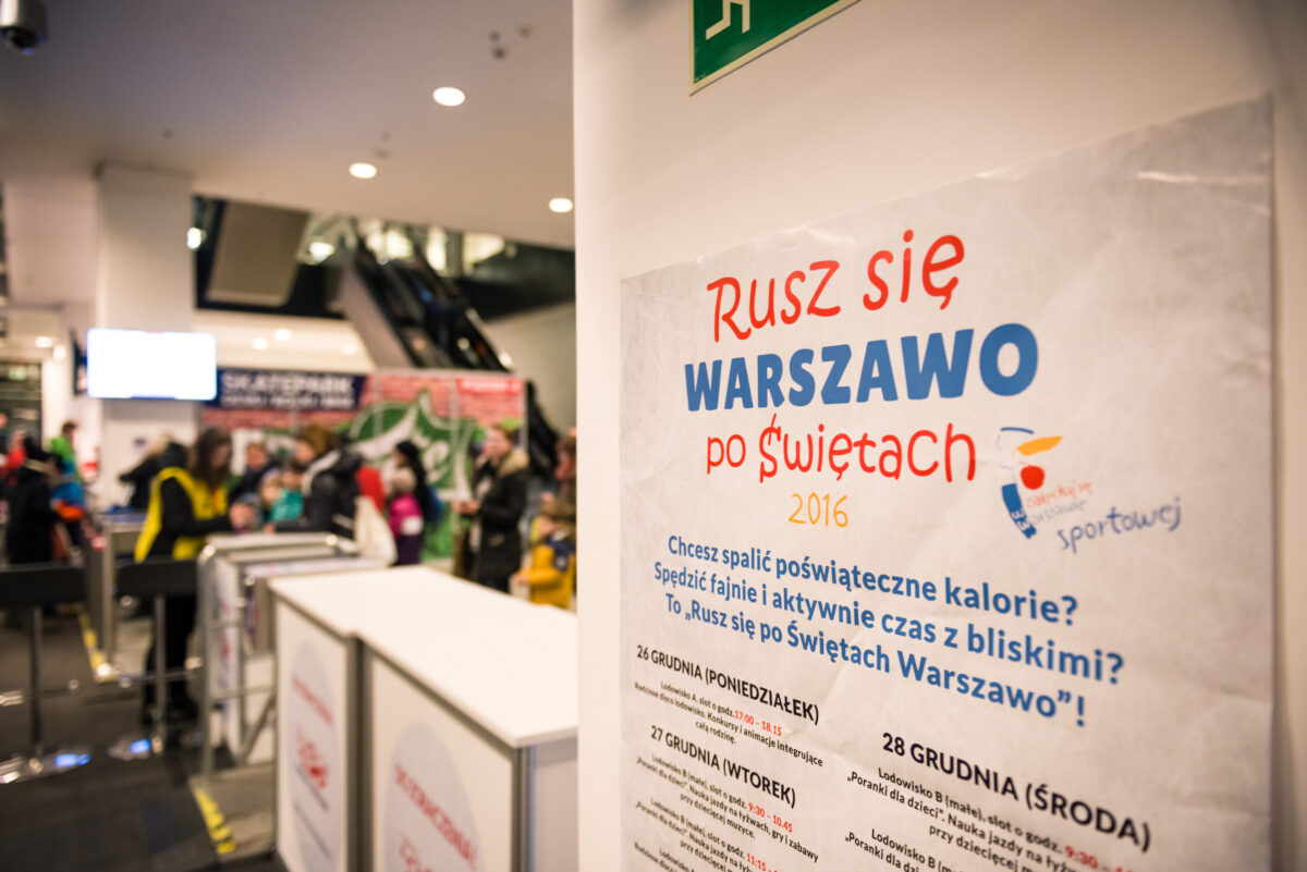 The poster, titled "Move Warsaw After Christmas 2016," is displayed in the foreground, with blurry figures in the background. The details of the event on the poster are written in Polish. Captured by a Warsaw event photographer, the setting appears to be an indoor public space with a modern interior.  