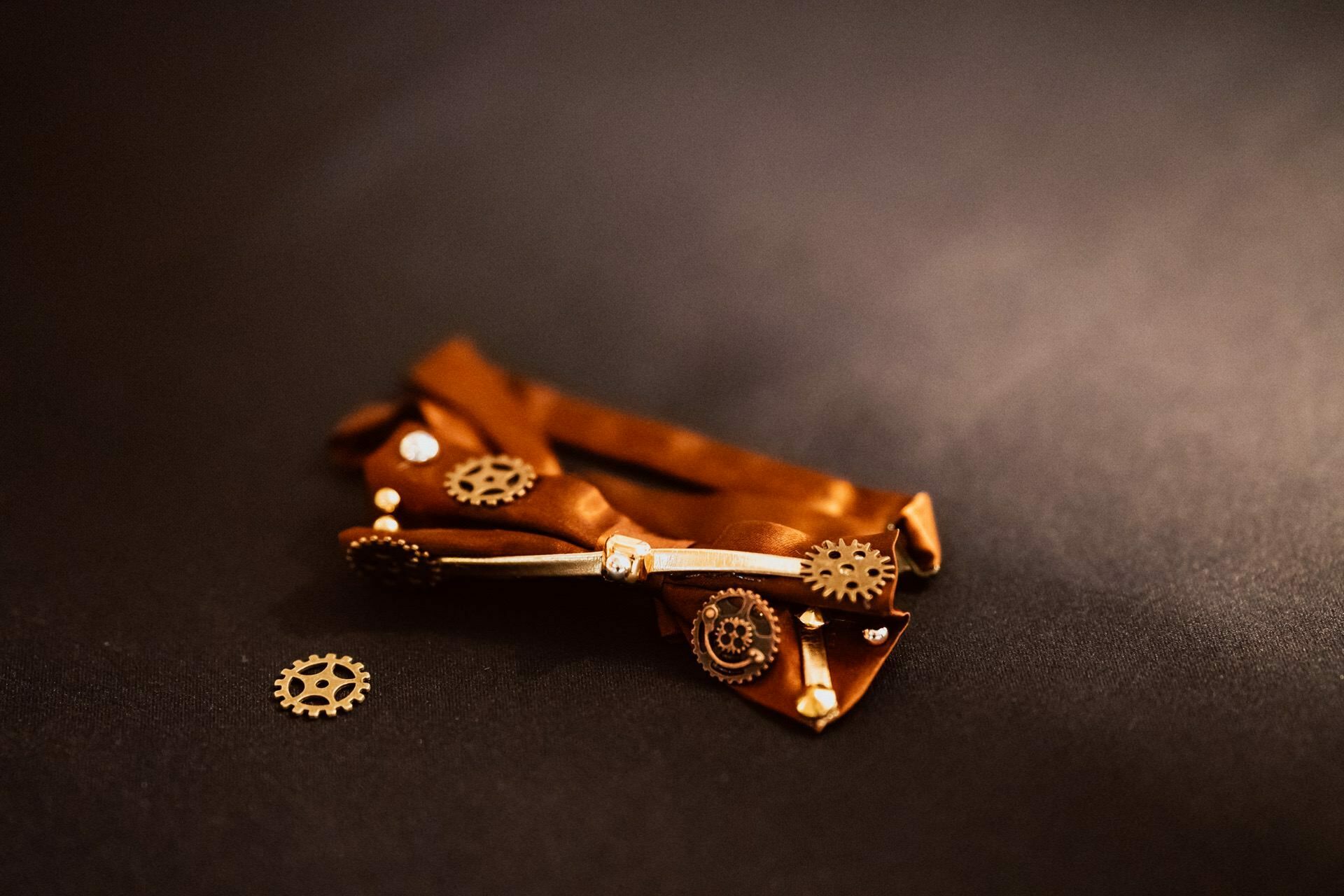 A handcrafted steampunk-style bow tie rests on a dark surface, decorated with small metal gears and mechanical parts. Nearby, loose, small hardware completes the intricate design, perfect for adding style to any photographic event. 