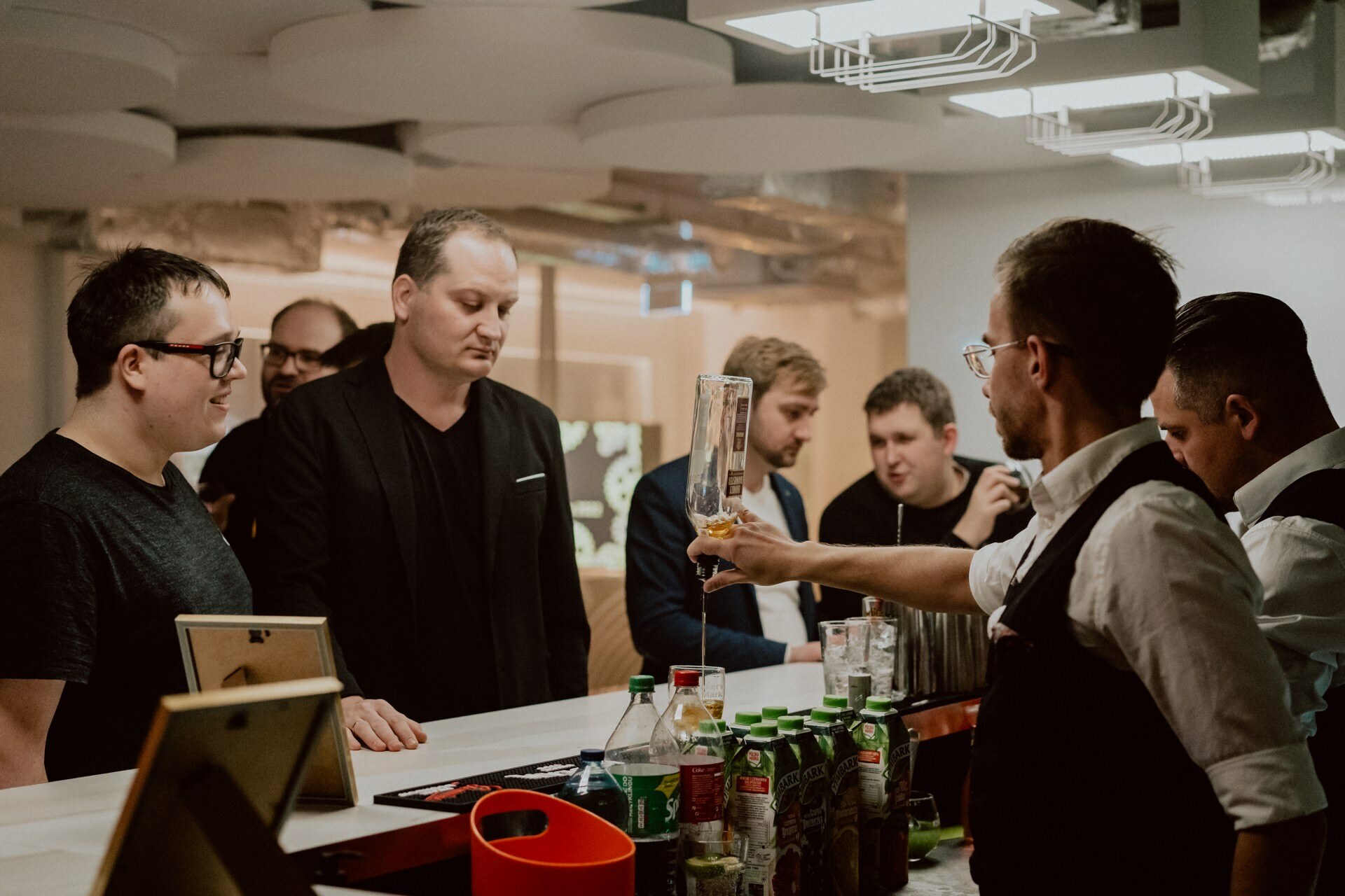 A bartender prepares drinks behind the bar for a group of men, casually chatting and watching the bartender work. Various bottles, mixers and drink ingredients are scattered on the countertop. The venue resembles a social gathering in a modern setting, making it ideal for event photography.  