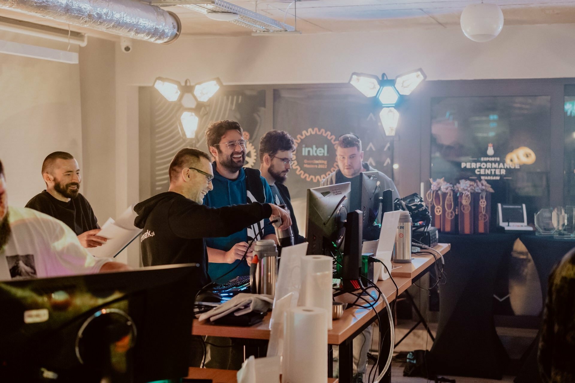 A group of people are gathered around computer monitors in a well-lit room. They seem engaged and smiling, suggesting an atmosphere of cooperation or celebration. In the background are illuminated signs and display cases that add to the professional setting. This vibrant scene could be part of a photo essay of a Marcin Krokowski event.   