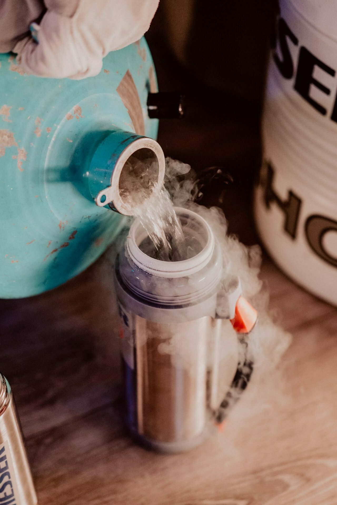 Close-up of a gloved hand pouring liquid nitrogen from a large insulated container into an open flask. Misty nitrogen vapor rises from the flask as the liquid is transferred. The scene takes place in a room on a wooden surface, which was perfectly captured by Marcin Krokowski in a photo report of the events.  