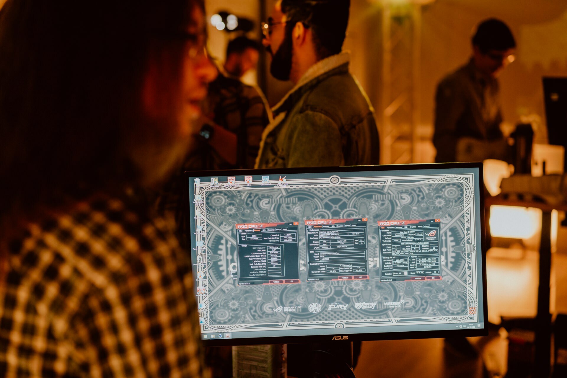 A computer monitor displays detailed settings, probably from software or a game, in a dimly lit room with many people doing activities. In the foreground you can see a person in a checkered shirt, out of focus, looking at the screen. This scene could be a fascinating photo essay of the event.  