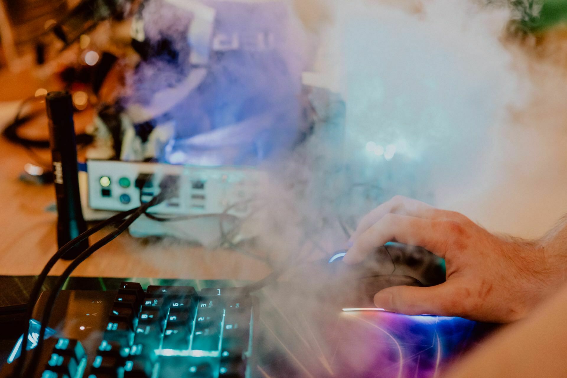 The hand of a person using a computer mouse next to a backlit keyboard with colorful keys. Nearby are various electronic devices and cables, partially obscured by a thick cloud of smoke or fog. This scene captures the essence of Marcin Krokowski's event photo report.  