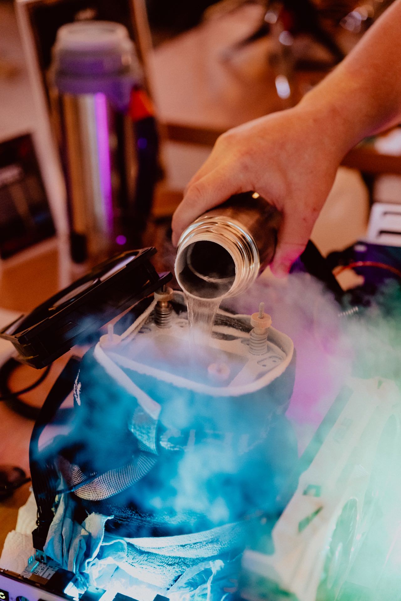 A hand pours liquid from a metal flask into a container emitting colored smoke. The setup appears to involve electronic equipment and suggests the use of a refrigerant, probably in a PC or technology device environment, as likely captured in the photo report of the event. 