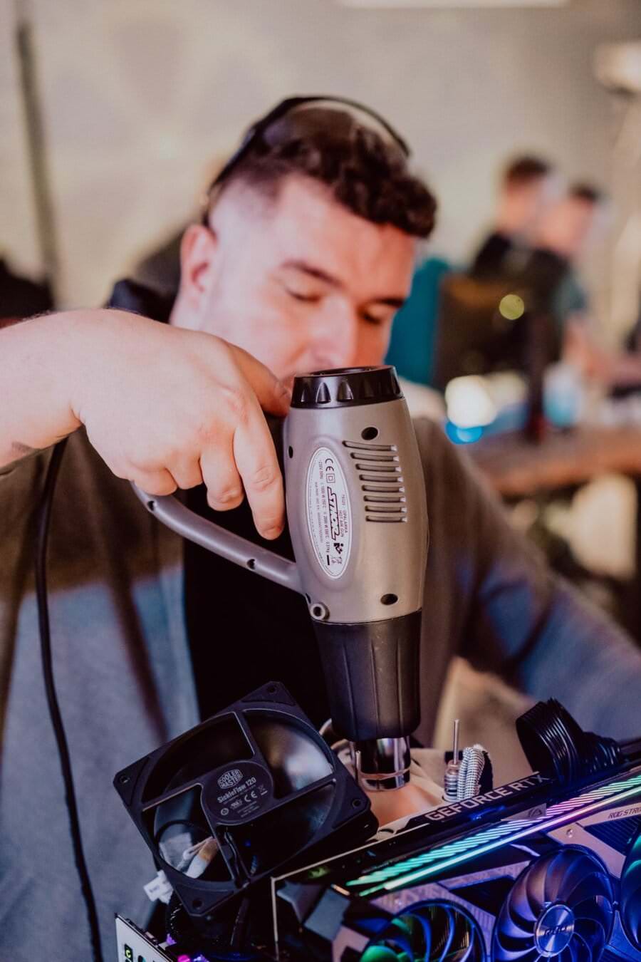 A person dressed in a gray hooded sweatshirt is using a tanning machine on a computer component with cooling fans attached. The background is blurred, suggesting a workshop or technological environment with other people and equipment, capturing the essence of the photo event. 