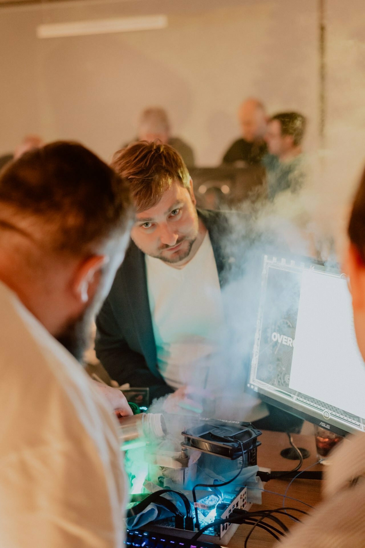 Marcin Krokowski stares intently at a computer screen amid a smoky environment. Surrounded by other people working on computers, various technical devices can be seen on a table. The scenery appears to be a collaborative or technology-based event, capturing the essence of the event photo report.  