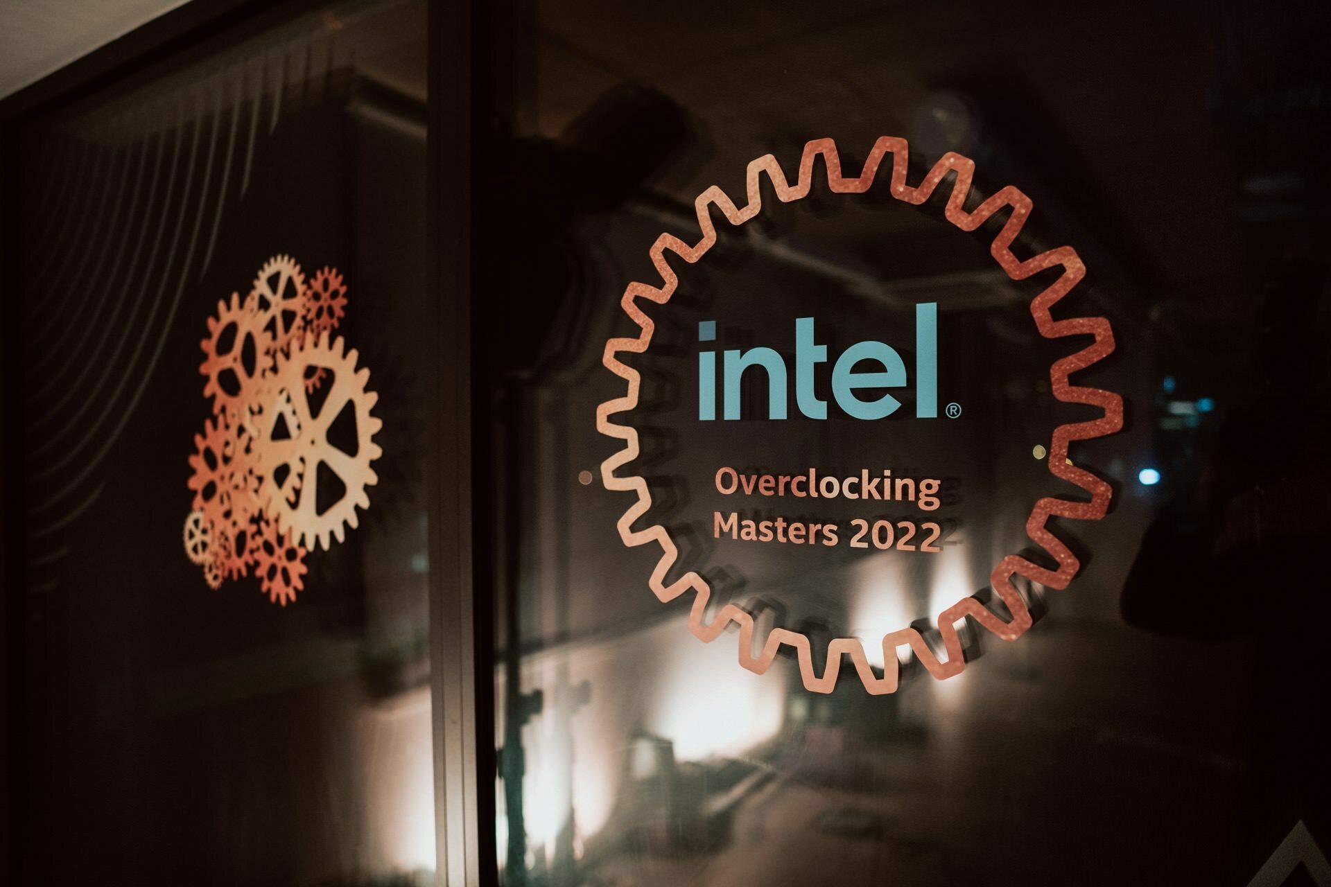 A glass panel with decorations of two gears and the words "Intel Overclocking Masters 2022" inside a large gear structure. The background is blurred and the ambient light creates a warm and inviting atmosphere, beautifully captured by the skills of event photographer Marcin Krokowski. 