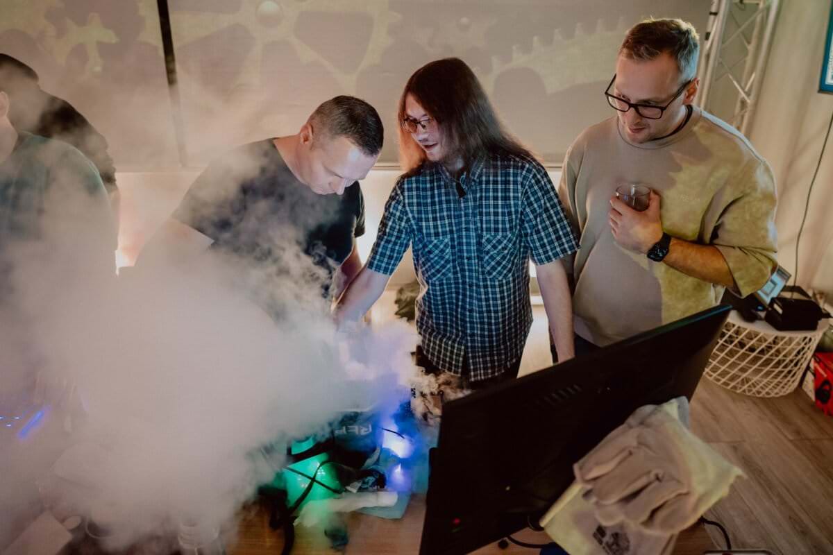 Three people stand around a table with a computer monitor, surrounded by fog or smoke. One person points to something on the table, while the other watches intently. The atmosphere seems focused and curious, and the equipment-like decorations in the background convey a detailed photo report of the event.  