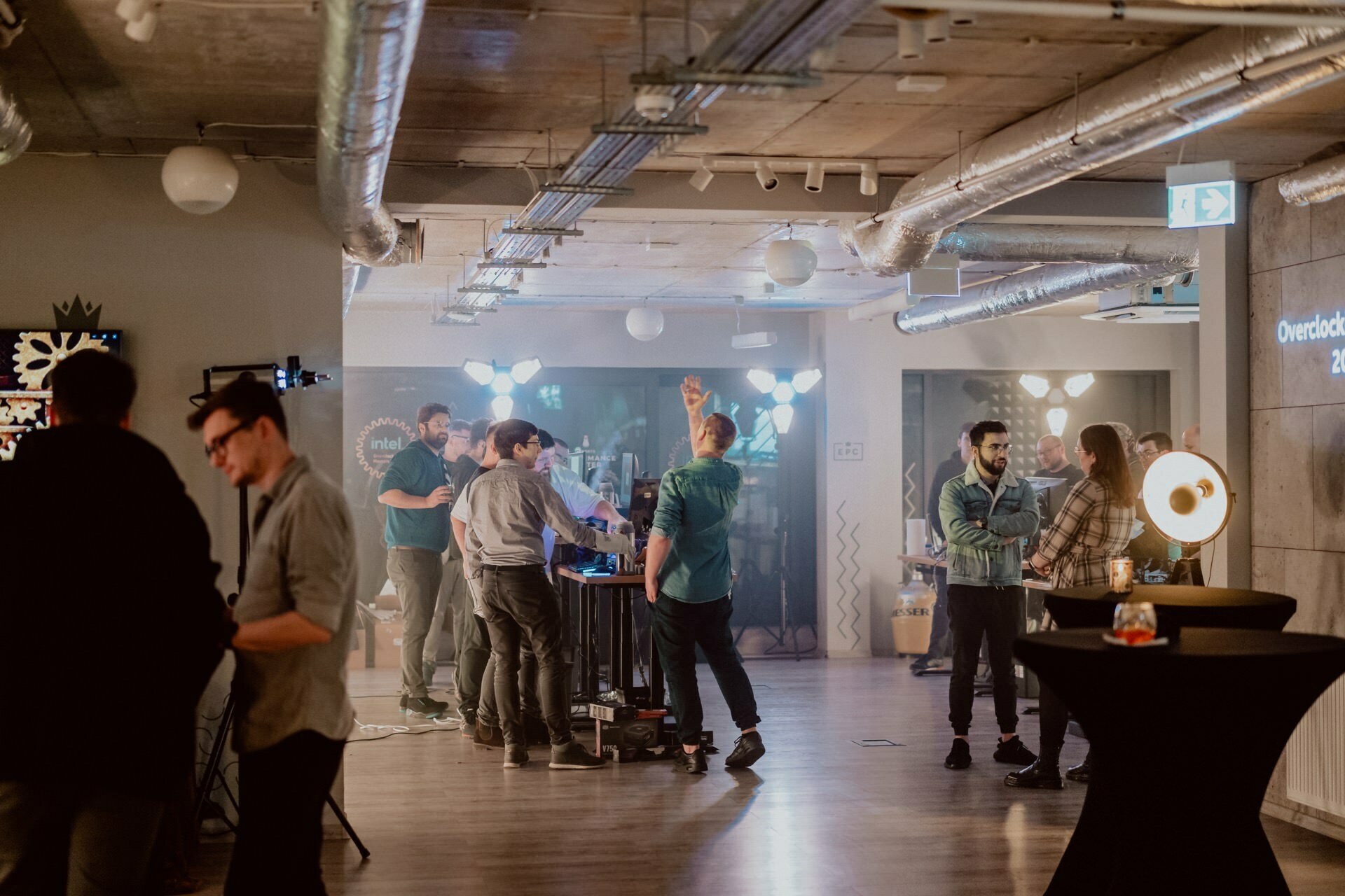 A group of people at a casual gathering in a room. Some are engaged in conversation, others are working on technical equipment. The room features an industrial decor with exposed pipes and ducts, while moody lighting creates a casual atmosphere. Perfect for photographer Marcin Krokowski to capture the moments.   