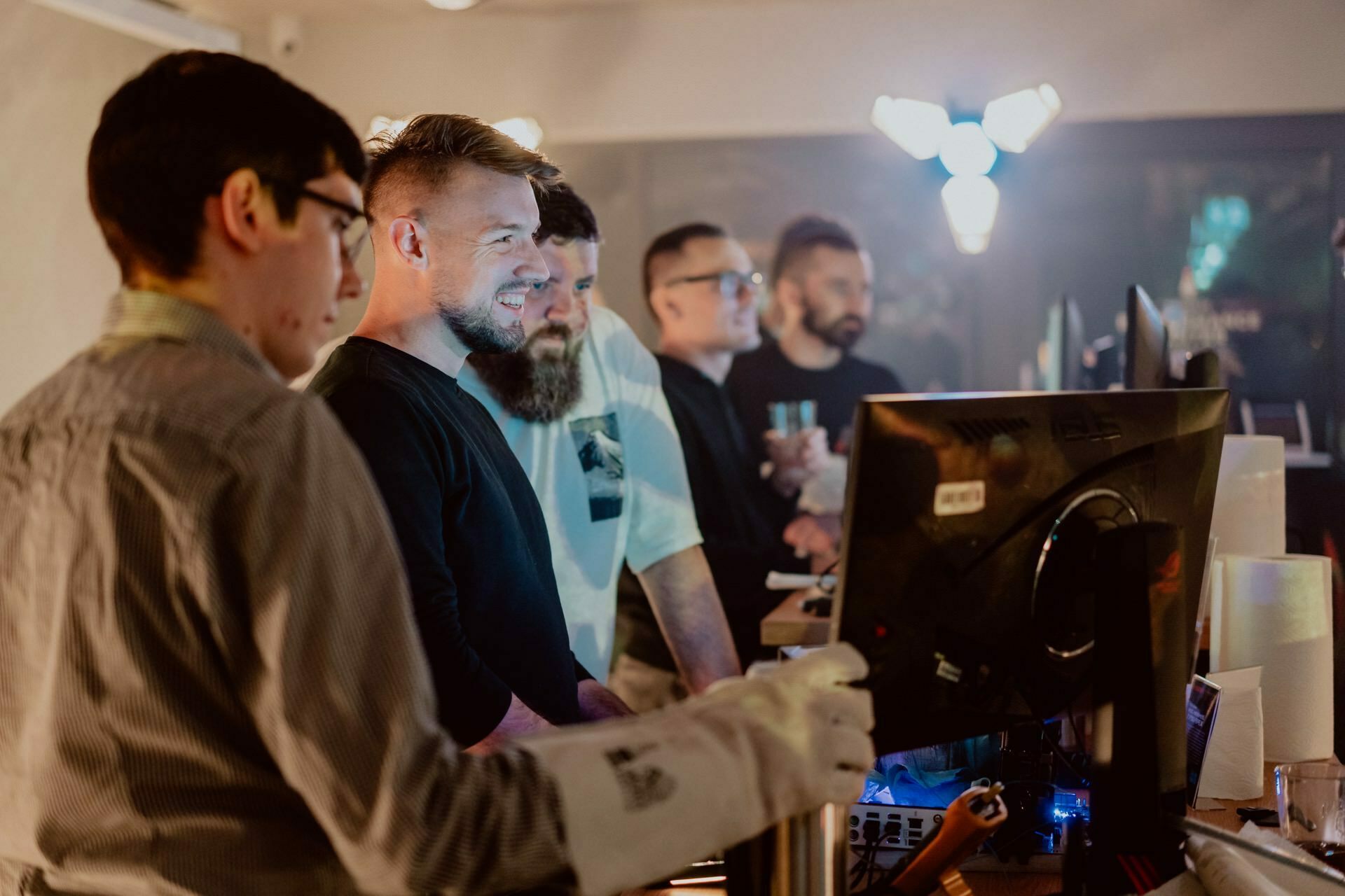 A group of five men work together in an office, clustered around a computer screen. Some are smiling as they discuss. One man is holding a drink, and another is wearing a pair of VR gloves. The room is softly lit, and its modern decor allows you to capture the moment like *event photography* by Marcin Krokowski.   