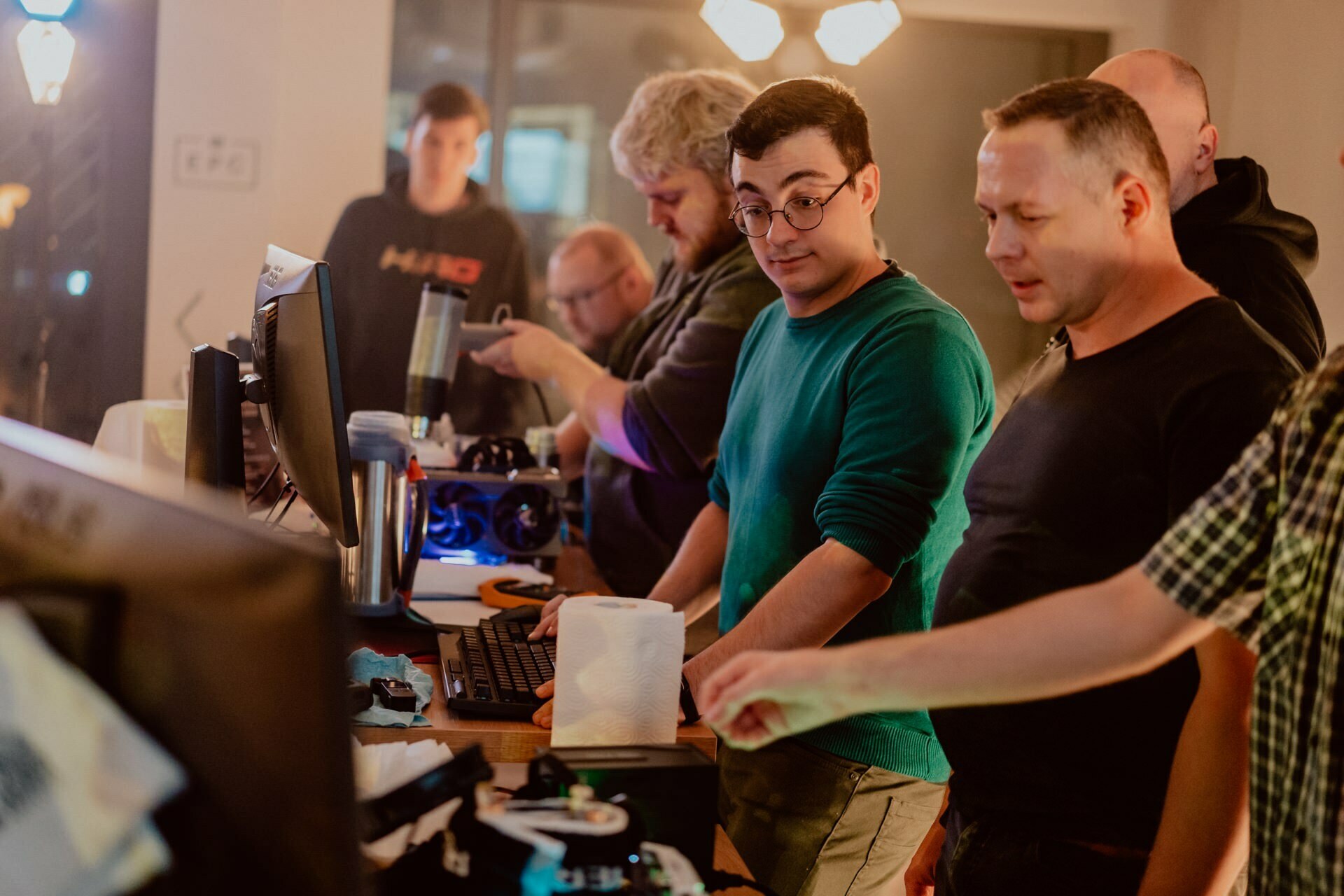 A group of people are gathered around a table with computers and electronic equipment, actively engaged in discussion. One man wearing glasses and a green sweater stares intently at the screen, while others around him point and talk. The scene is brightly lit, and captures an engaging *photography of the event* by Marcin Krokowski.  