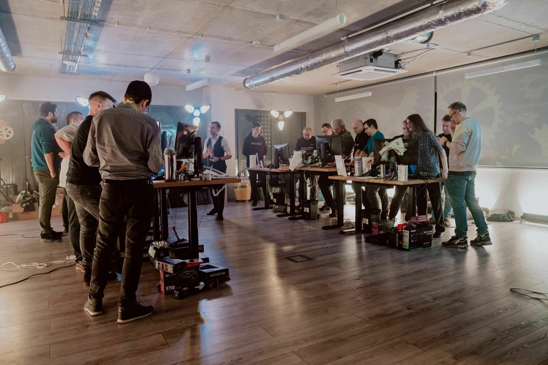 A group of people gathered in an industrial-style room with concrete ceilings, working together around standing desks with computers. The room features modern lighting and exposed air ducts. Some chatted while others focused on screens, as recalled in a photo report of the event by marcin krokowski.  