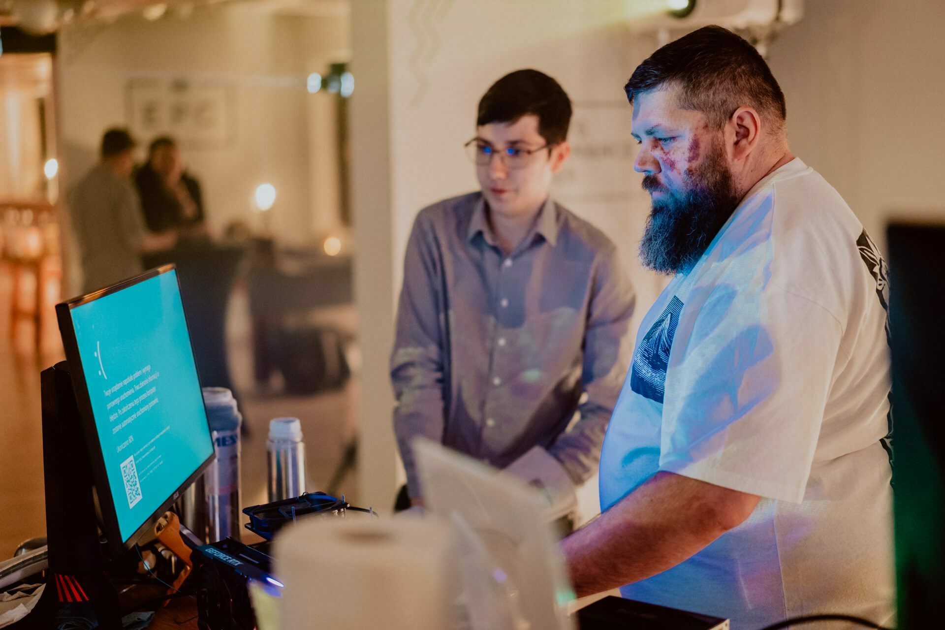 Two men are setting up music equipment during an event taking place in a room. A bearded man in a white t-shirt operates a laptop, while another man in glasses and a button-down shirt observes. The background is slightly out of focus, with other people and decorations visible, perfect for a photo essay on events by Marcin Krokowski.  